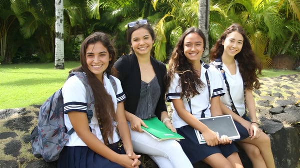Cuatro estudiantes de la Sacred Hearts Academy sonríen y posan juntas en un jardín tropical con palmeras.