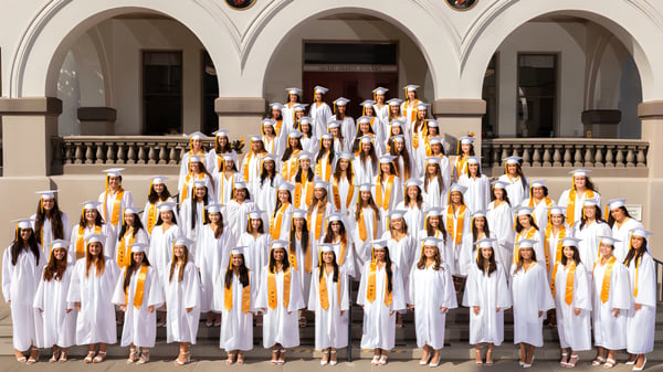 Estudiantes de la Sacred Hearts Academy están de pie en túnicas blancas con cintas amarillas frente a un edificio decorado con columnas.