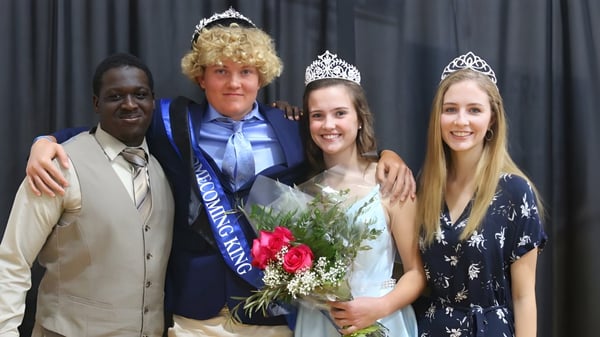 Una alumna de la Saint Francis Catholic Academy lleva una corona y sostiene un ramo de flores frente a un telón oscuro.
