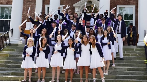 Un gran grupo de graduados está de pie en túnicas blancas y moradas en los escalones de un edificio de ladrillo de la Saint James School.