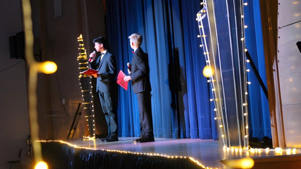 Dos personas en ropa formal están en el escenario de la Saint-Lambert International High School frente a una cortina azul con luces.