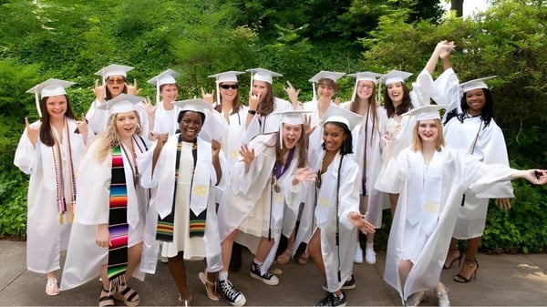 Un grupo de alumnas de la Salem Academy está reunido en el área exterior verde con togas de graduación blancas.