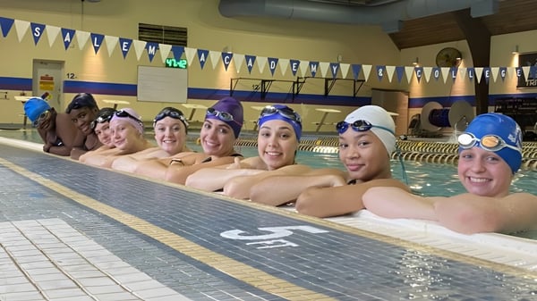Un grupo de alumnas sonrientes de la Salem Academy está sentada al borde de una piscina cubierta brillante con decoración azul y blanca.