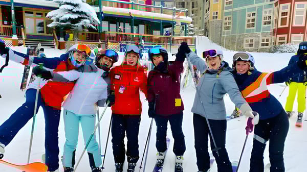 Un grupo de estudiantes de la Salmon Arm Secondary está en colorido equipo de esquí en un paisaje nevado frente a edificios y un árbol cubierto de nieve.