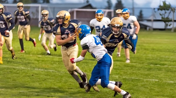 Un grupo de jugadores de fútbol de la Salmon Arm Secondary juega intensamente en el campo de césped con espectadores al fondo.