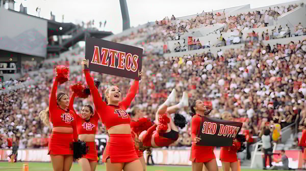 Estudiantes de la San Diego State University animan en el estadio con uniformes rojos y trajes de animación.