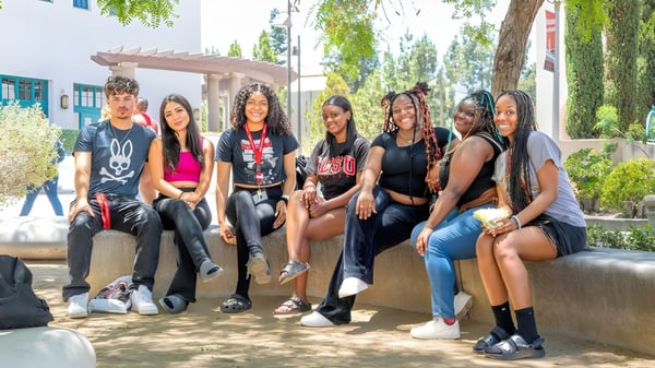 Un grupo de estudiantes está sentado juntos en un banco en el área exterior del campus de la San Diego State University.