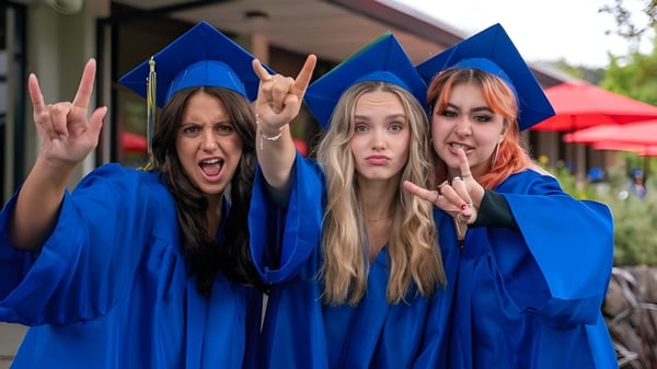 Tres estudiantes de la San Domenico School llevan togas de graduación azules y posan frente a una carpa roja con signos de paz.