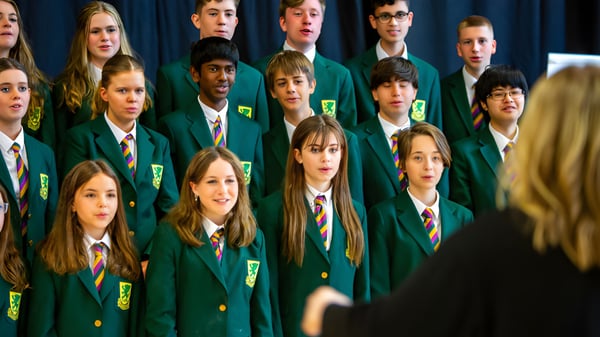 Un grupo de estudiantes en uniformes escolares verdes está en filas frente a un fondo oscuro en el campus de la Sandford Park School.