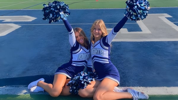 Dos cheerleaders realizan una coreografía en el campo deportivo del Santa Barbara Unified School District con uniformes azules y blancos.