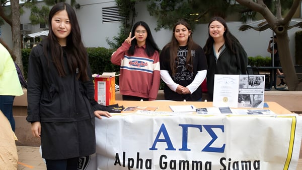 Cuatro estudiantes están detrás de una mesa con un banner de Alpha Gamma Sigma en el campus del Santa Monica College.