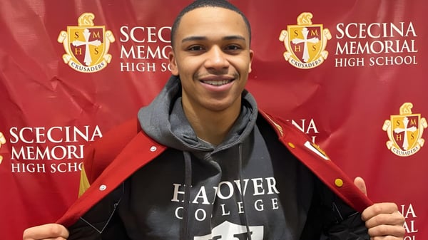 Un estudiante está de pie frente al logo de la Scecina Memorial High School sobre un fondo rojo.