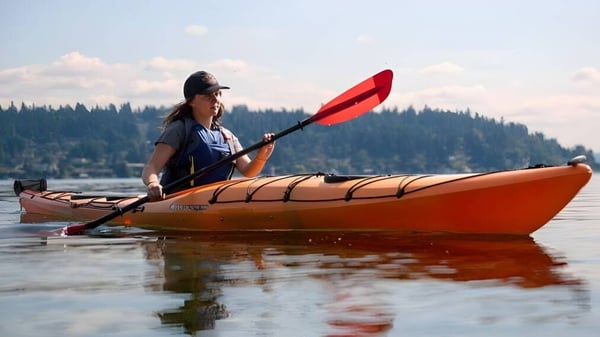 Una persona rema en un kayak en un lago tranquilo frente a montañas boscosas cerca del Schull Community College.