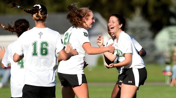 Estudiantes de Scoil Aireagail celebran juntos en el campo de deportes con árboles y un cielo claro de fondo.