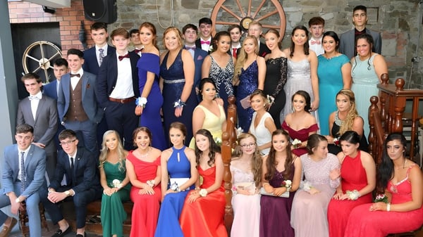 Un grupo de estudiantes vestidos formalmente en el campus de la Scoil Mhuire Kanturk frente a un fondo rústico con una rueda de madera.