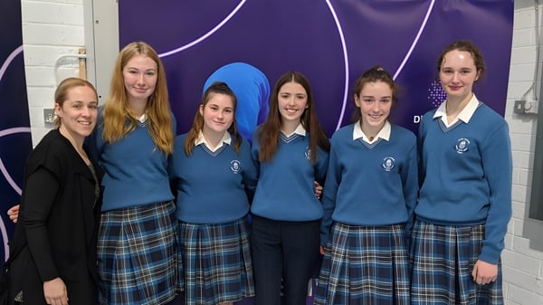 Un grupo de alumnas de la Scoil Mhuire (Trim) están frente a un fondo púrpura con patrones abstractos.