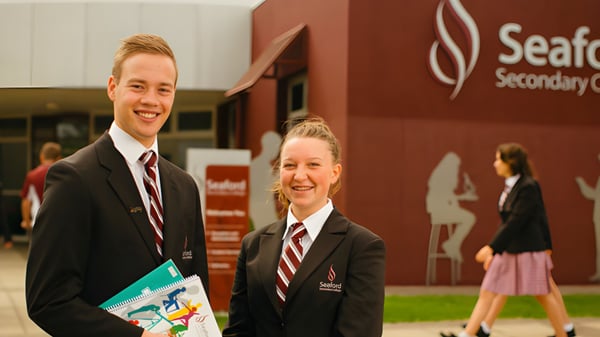 Dos estudiantes en uniformes escolares están frente al cartel del Seaford Secondary College sosteniendo un documento.