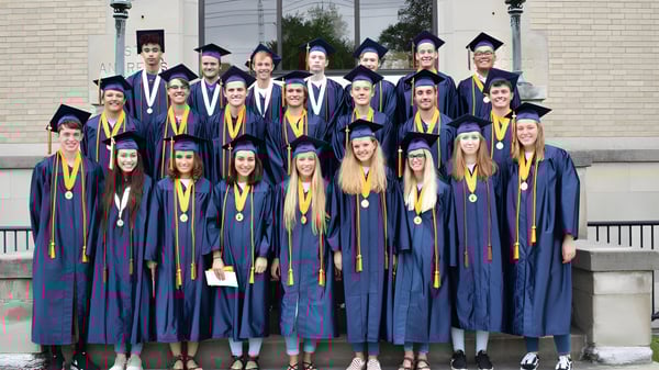 Graduados de la Seton Catholic High School están de pie en togas de graduación azules y amarillas frente a un edificio de ladrillo.