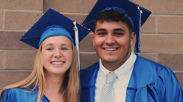 Dos graduados sonrientes de la Shore Regional High School en togas de graduación azules están frente a una pared de ladrillo.