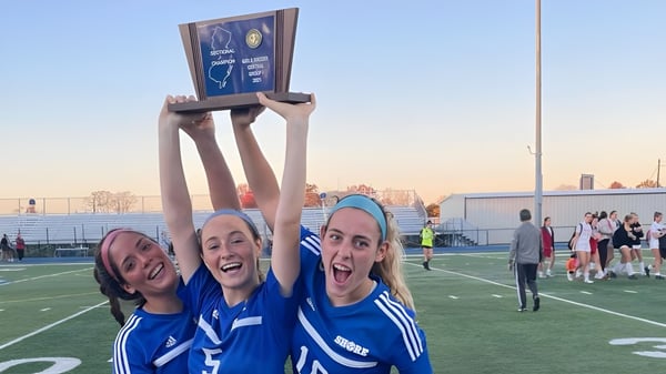 Tres alumnas de la Shore Regional High School celebran con un trofeo en el campo deportivo frente a un paisaje urbano.