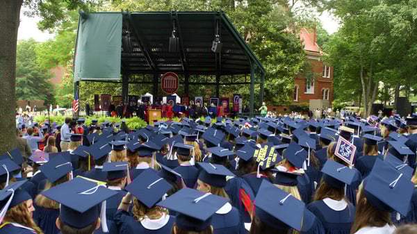 Los graduados de la Sidcot School se reúnen al aire libre en togas y birretes azules durante la ceremonia de graduación frente a un escenario.