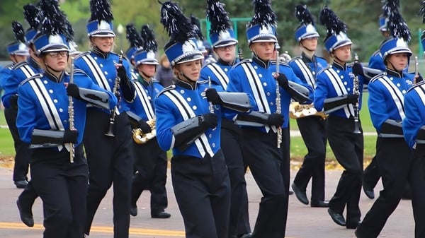 Miembros de la banda marcial de Sioux Falls Christian School están en uniforme azul y negro sobre un césped con árboles al fondo.