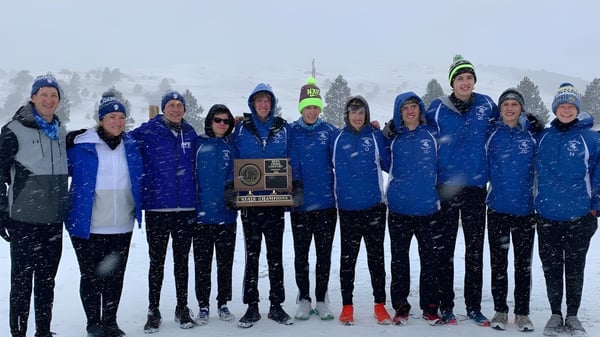 Un grupo de alumnas y alumnos de Sioux Falls Christian School está en uniformes de deportes de invierno azules y grises frente a un paisaje montañoso nevado.
