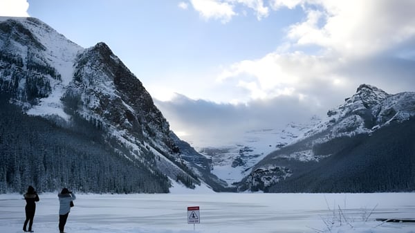 Dos personas pasean junto a un lago congelado con montañas nevadas al fondo cerca de la Sir Guy Carleton Secondary School.