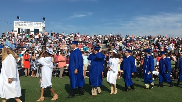 Un gran grupo de graduados de la Smithfield State High School está de pie en un prado con sus togas y birretes bajo un cielo azul.