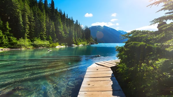 Un muelle de madera se extiende sobre un lago claro cerca de la South Delta Secondary School con montañas y bosques en el fondo.