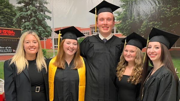 Un grupo de cinco graduados está frente al edificio de la South Grenville District High School celebrando su graduación.
