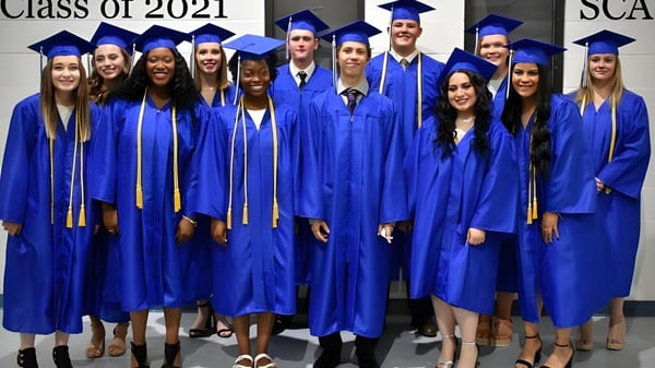 Un grupo de graduados de la Southwest Christian Academy posan con togas azules frente a una pared con la inscripción Class of 2021.