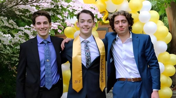 Tres estudiantes en ropa formal están de pie frente a árboles en flor y globos amarillos en el campus de la Spectrum Community Secondary School.