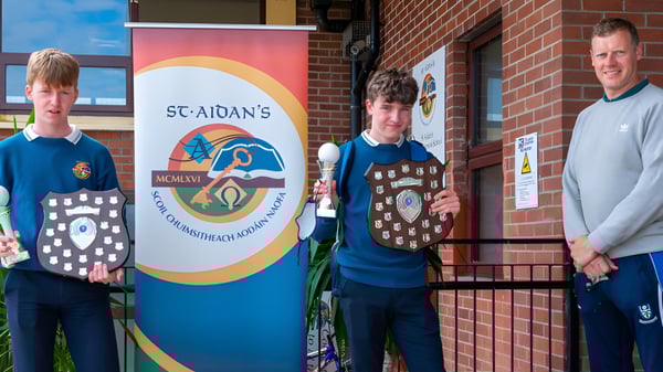 Tres alumnos de St. Aidan's Comprehensive School están frente a un edificio de ladrillo con un gran cartel de la escuela.
