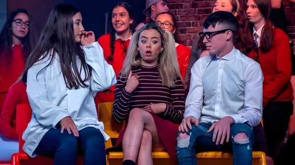 Un grupo de alumnos de St. Aidan's Comprehensive School está sentado en el escenario frente a una pared de ladrillo en una situación de actuación.