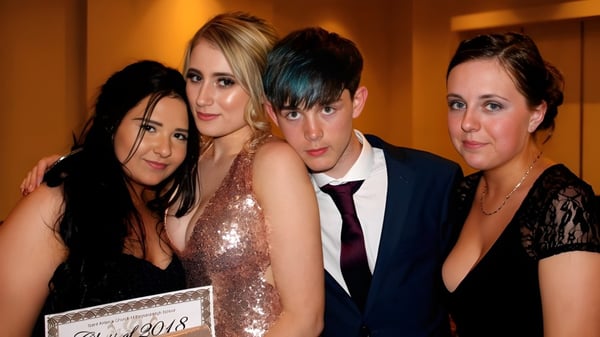 Cuatro personas vestidas de manera formal posan juntas frente a un lugar de eventos en el campus de St. Aidan's Comprehensive School.