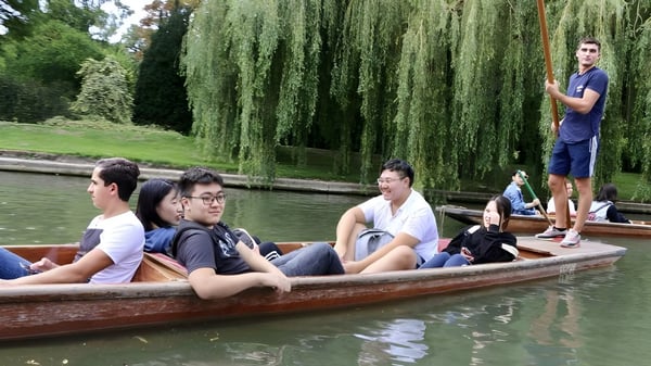 Un grupo de estudiantes del St. Andrew’s College se relaja en un bote de madera en un estanque con sauces.