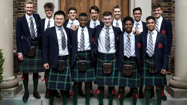 Un grupo de estudiantes en ropa formal está frente a una puerta de madera en el terreno del St. Andrew’s College.