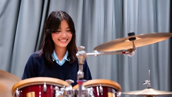 Una estudiante de la St. Catherine's School toca la batería con entusiasmo en un escenario con una cortina gris de fondo.