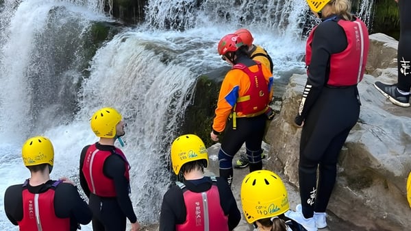 Un grupo de alumnas y alumnos de la St. Clare’s School están en trajes de protección coloridos frente a una cascada rugiente.