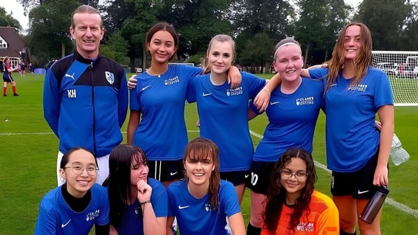 Un grupo de alumnas de la St. Clare’s School en trajes deportivos azules están de pie y sentadas en un campo de deporte con hierba y árboles.