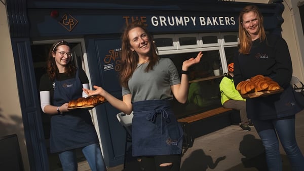 Tres alumnas del St. Colman’s Community College están frente a la tienda The Grumpy Bakers sosteniendo productos recién horneados.