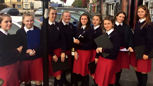 Alumnas de St. Dominic’s College están de pie en uniforme escolar en una escena de calle con edificios y vehículos de fondo.