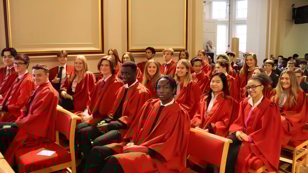Estudiantes de la St. Edmunds School están sentados en una ceremonia de graduación solemne en una sala con grandes ventanas y decoración lujosa.