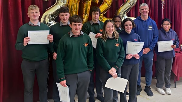 Estudiantes de St. Fergal’s College están en uniforme escolar con certificados frente a un fondo decorado en oro y púrpura.