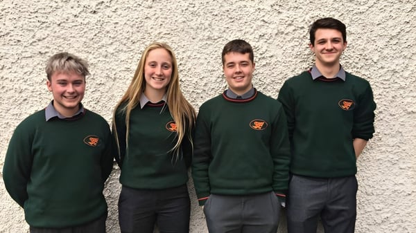 Cuatro estudiantes están de pie frente a una pared blanca con suéteres verdes en el campus de St. Fergal’s College.