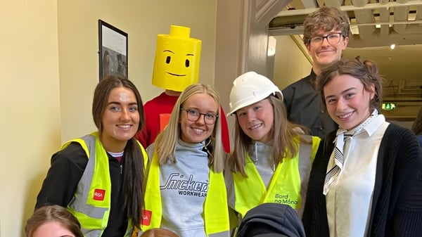 Un grupo de alumnas y alumnos del St. Finian’s College en chalecos de seguridad y cascos posan frente a una figura de dibujos animados amarilla.