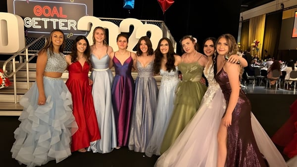 Un grupo de jóvenes en vestidos de gala formales se encuentra frente a un fondo con la inscripción GOAL GETTER en el campus de la St. Francis High School.