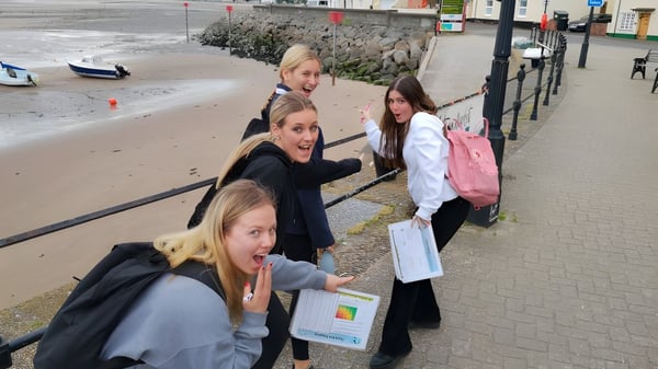 Un grupo de alumnas de St. George's Ascot está juntas en una acera cerca de un cuerpo de agua con costa rocosa y edificios al fondo.