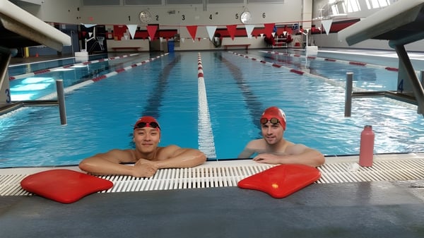 Dos estudiantes con gorros de baño rojos están sentadas sobre tablas de natación rojas en la piscina de St. George’s School.